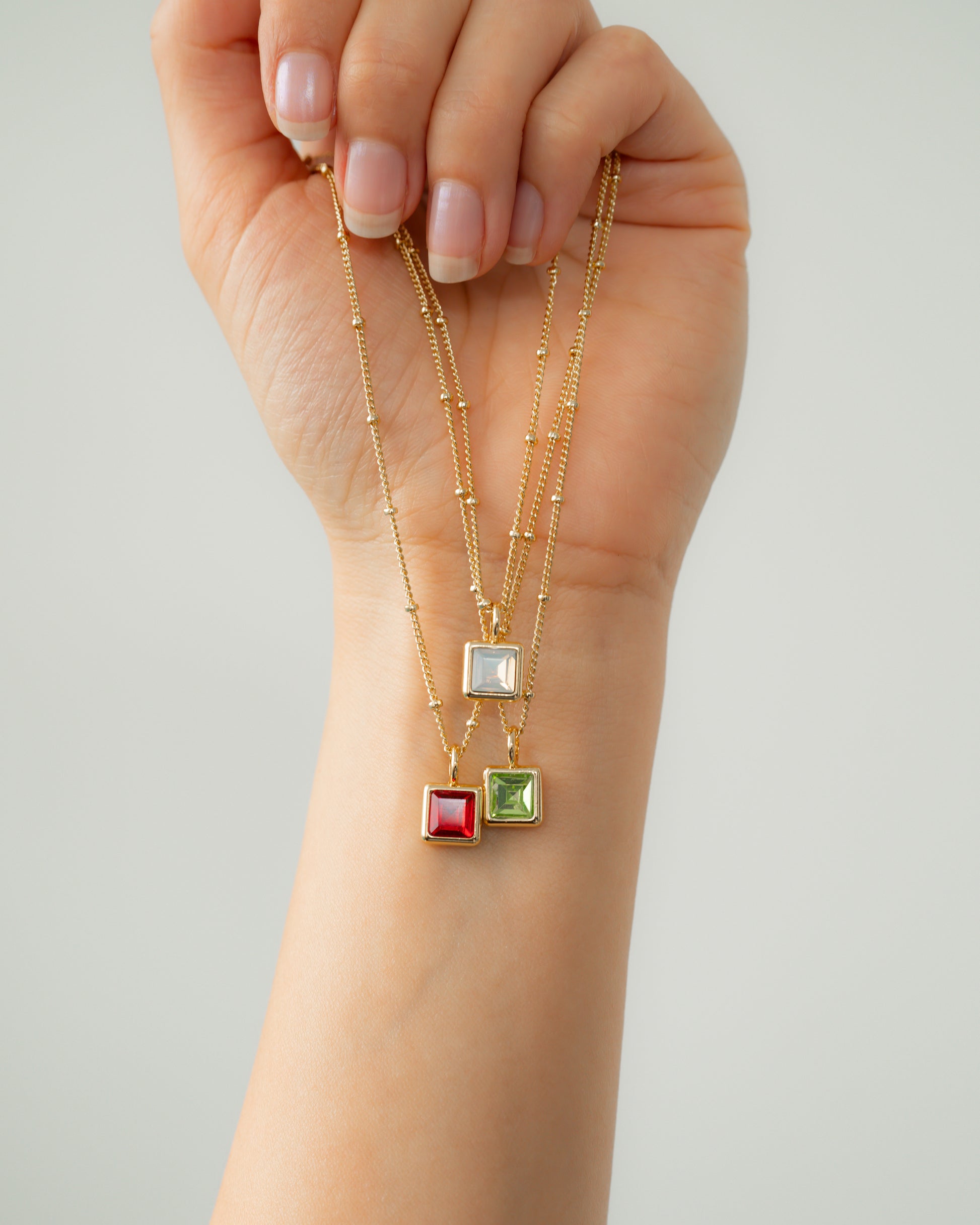 A hand holds three gold necklaces with square pendants of white, red, and green.