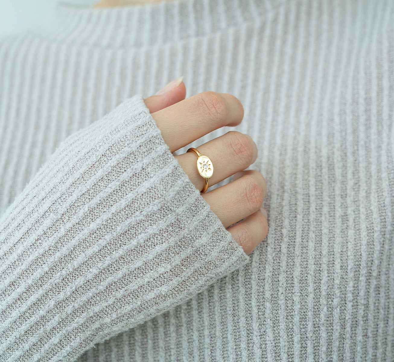 A person's hand wearing a gold ring with a starburst design on it rests against a light gray sweater.