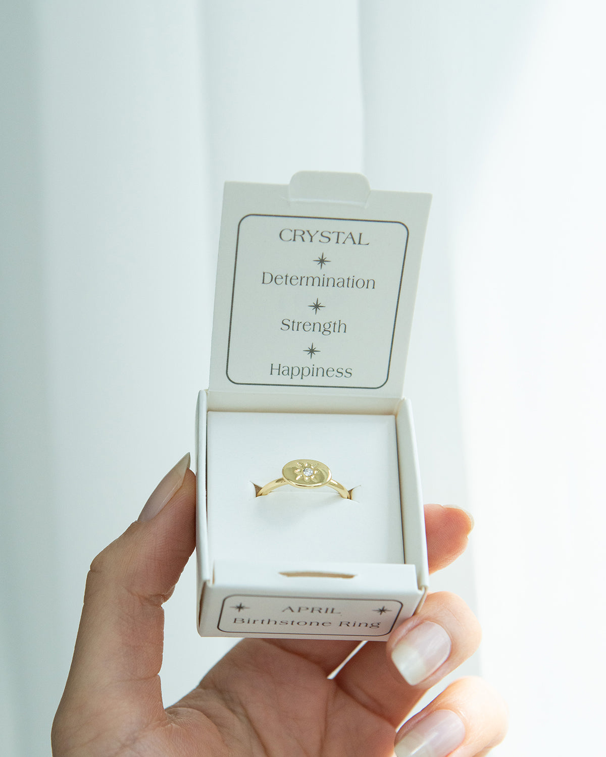 A hand holds a box with an "April Birthstone Ring" inside.