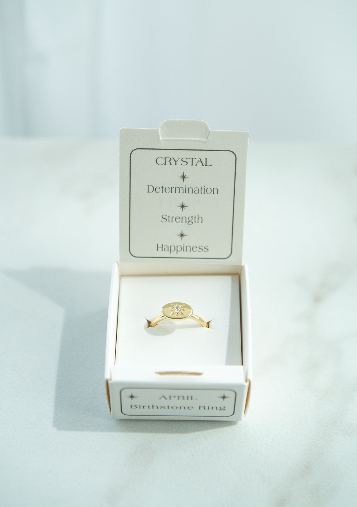 A gold "April Birthstone Ring" with a crystal sits inside its labeled white box.