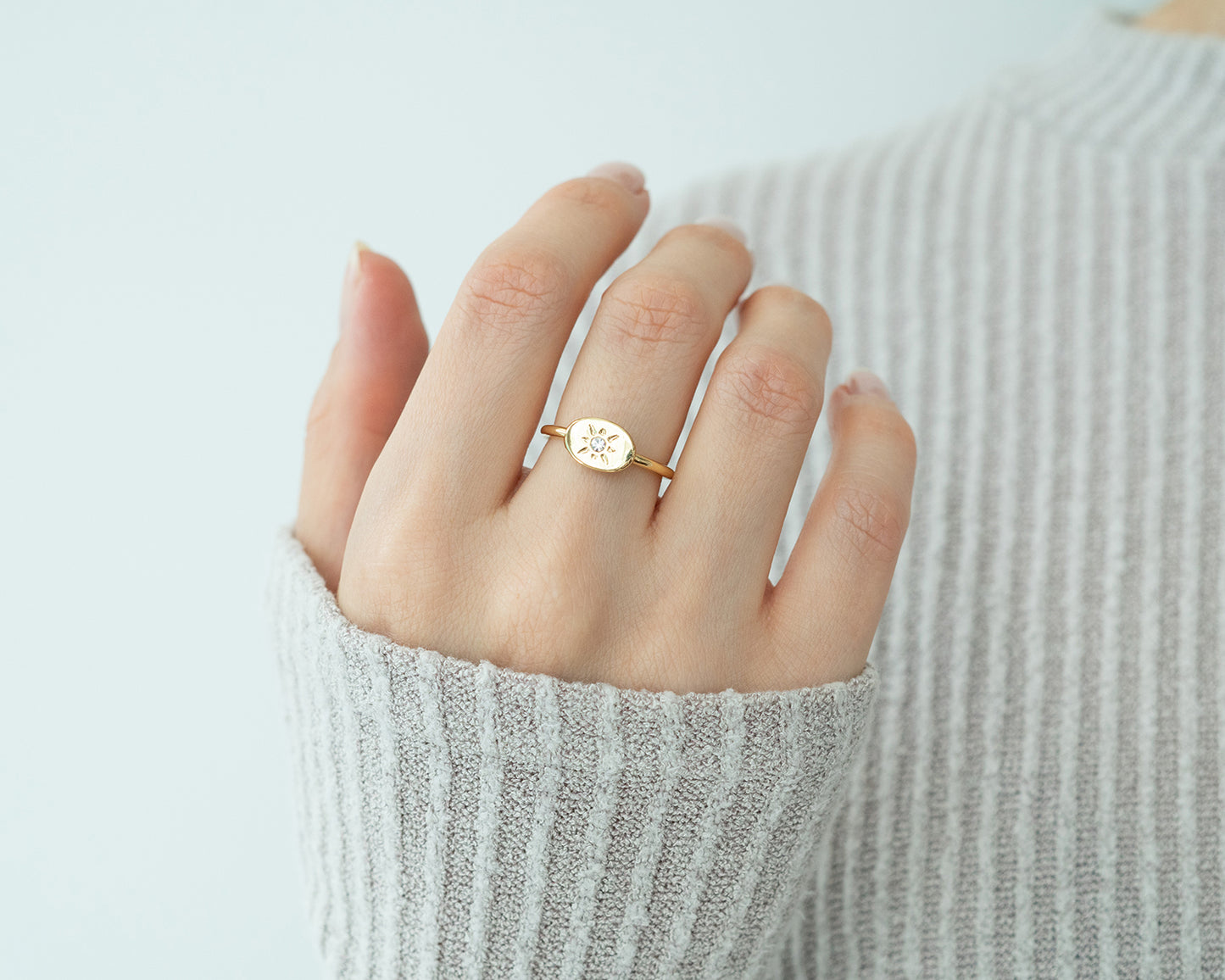 A hand wearing a gold ring with a sunburst design is posed in front of a gray sweater.