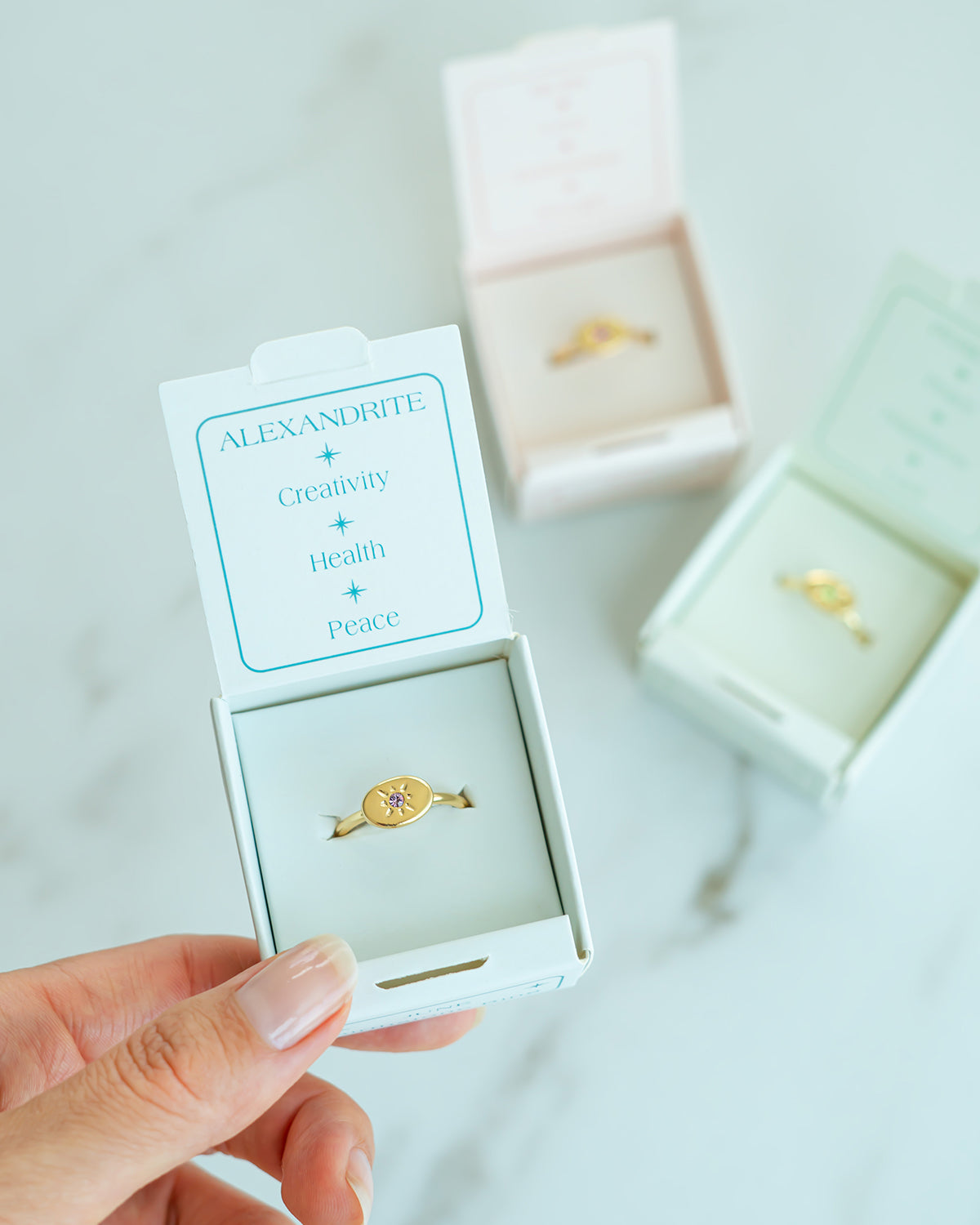 Hand holding an open jewelry box containing a gold ring with a purple stone, with other rings in similar boxes in the background.