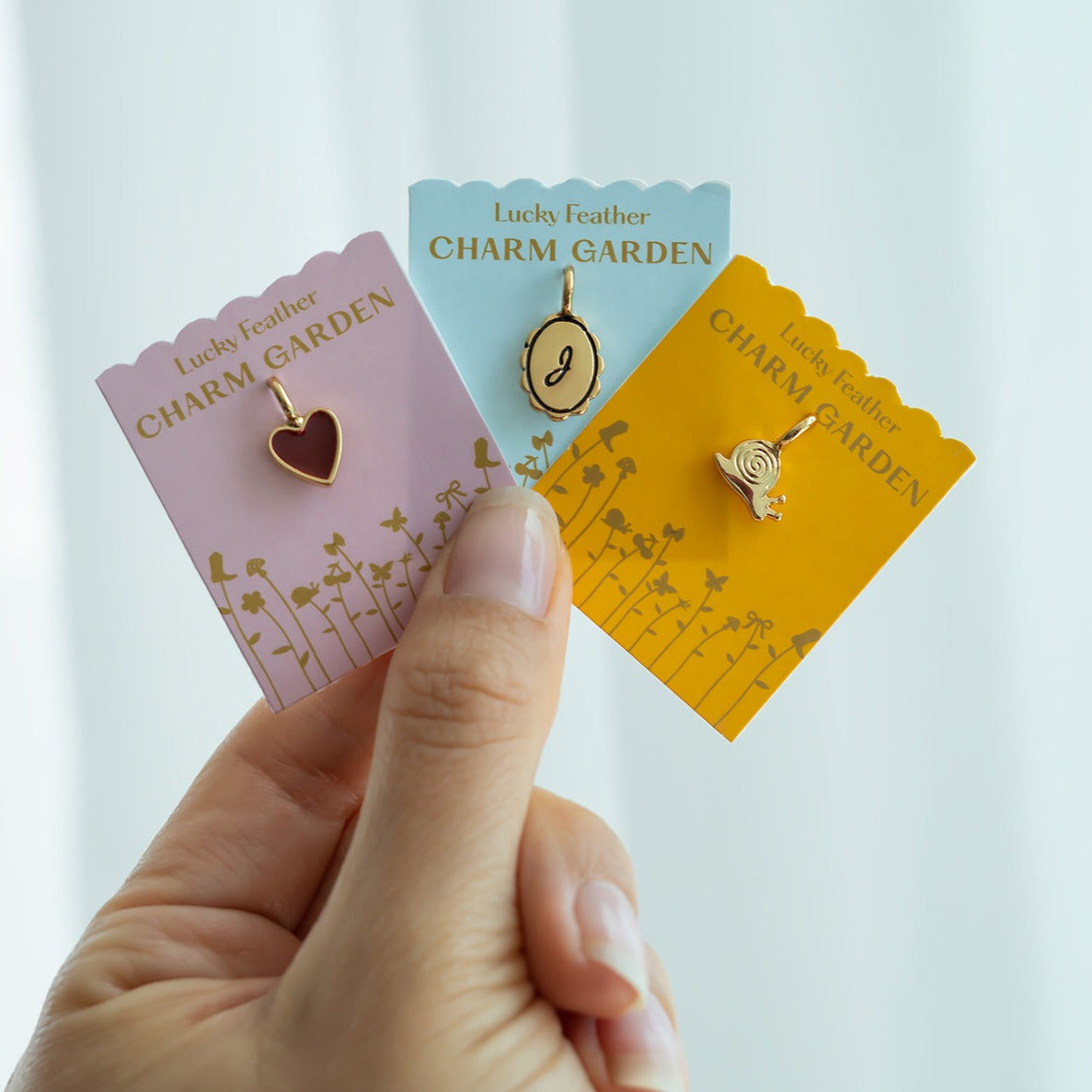 Hand holding three "Lucky Feather Charm Garden" cards with a heart, letter, and snail charm.