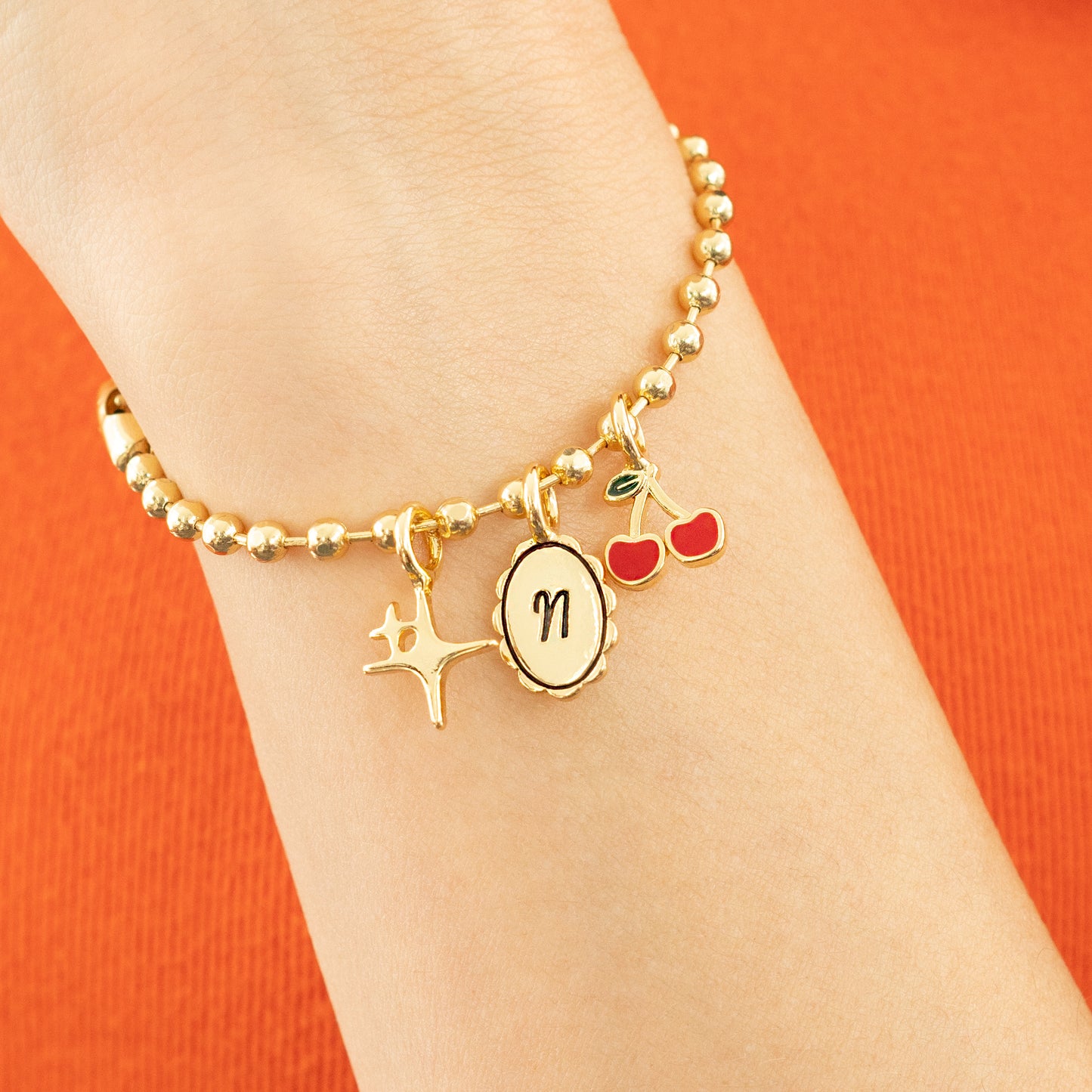 A wrist wearing a gold bead bracelet with star, initial, and cherry charms is shown against an orange background.