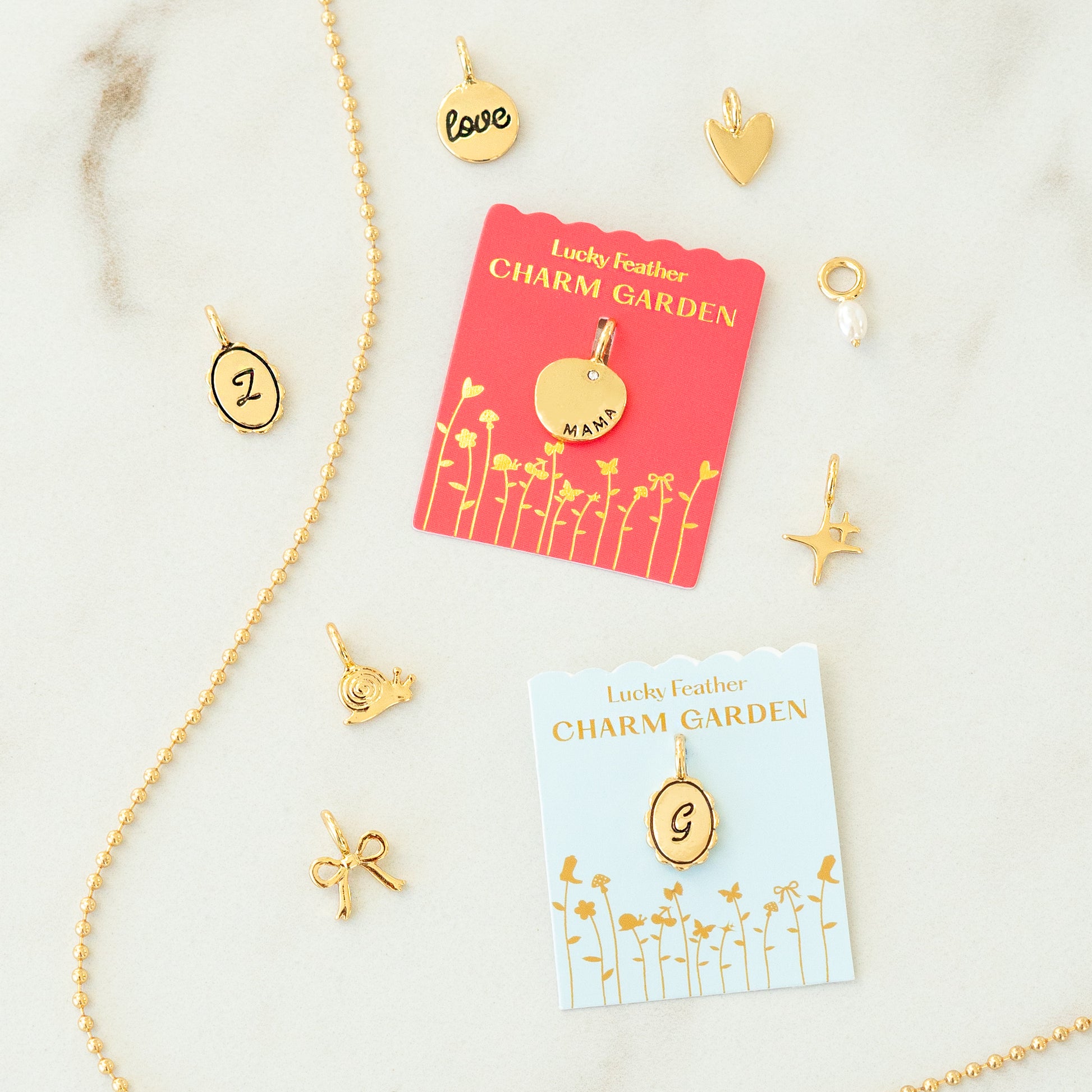 A selection of gold charms and a beaded chain, including two 'Lucky Feather Charm Garden' cards, on a marble-like surface.