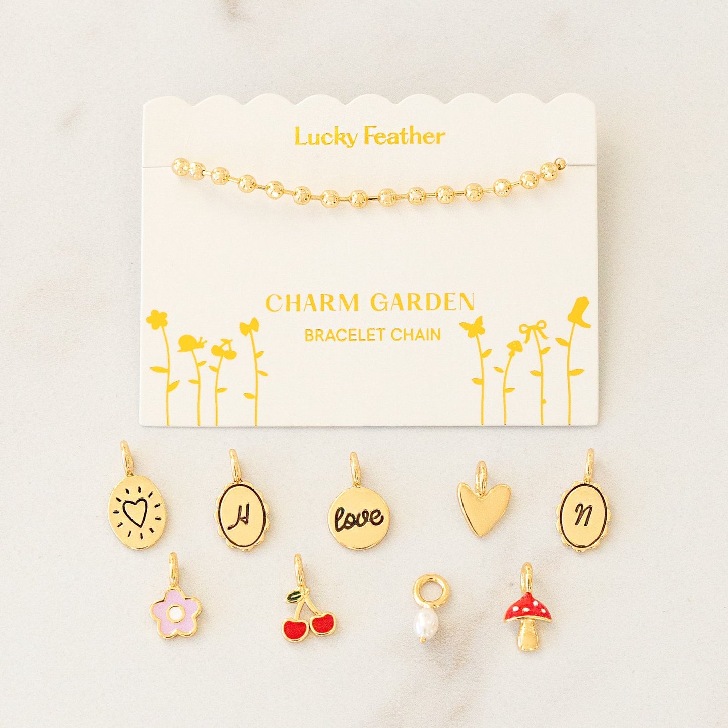 A gold ball chain bracelet displayed on a card labeled "Lucky Feather Charm Garden Bracelet Chain" with assorted gold charms scattered below.