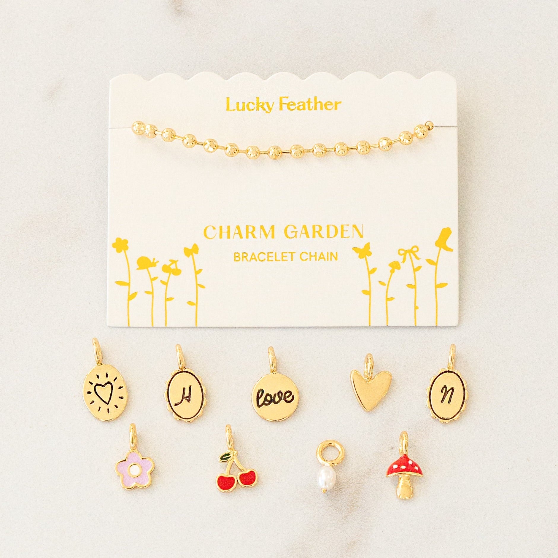 A gold ball chain bracelet displayed on a card labeled 'Lucky Feather Charm Garden Bracelet Chain' with assorted gold charms scattered below.