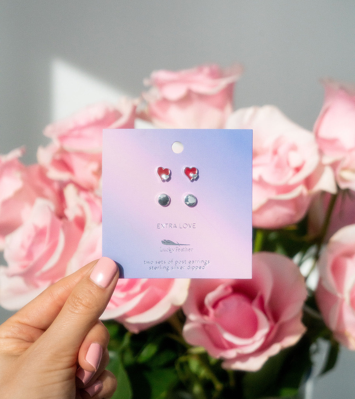 A hand holds a card of earrings in front of a blurry bouquet of pink roses.