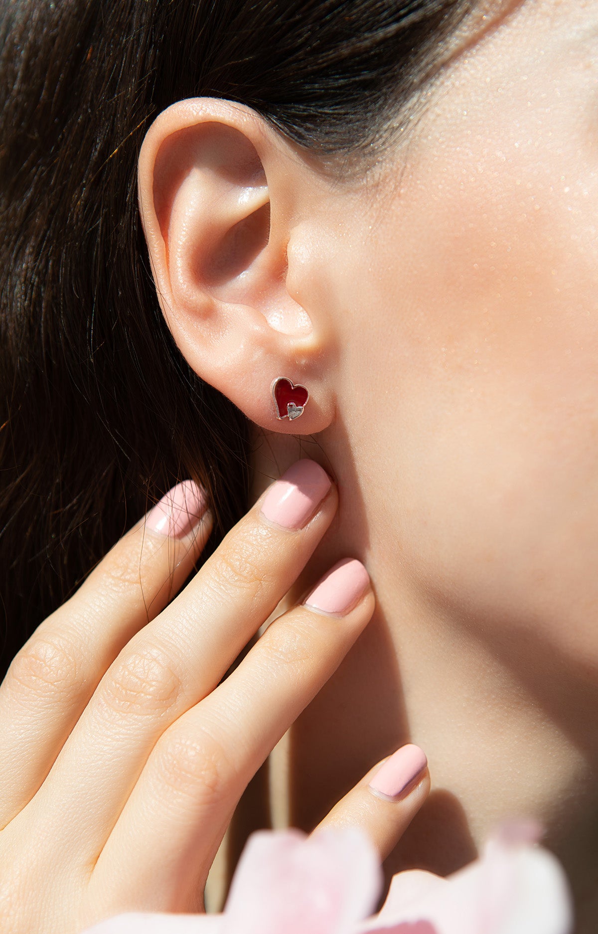 Close-up of a woman's ear with a red heart earring, her hand near her face.