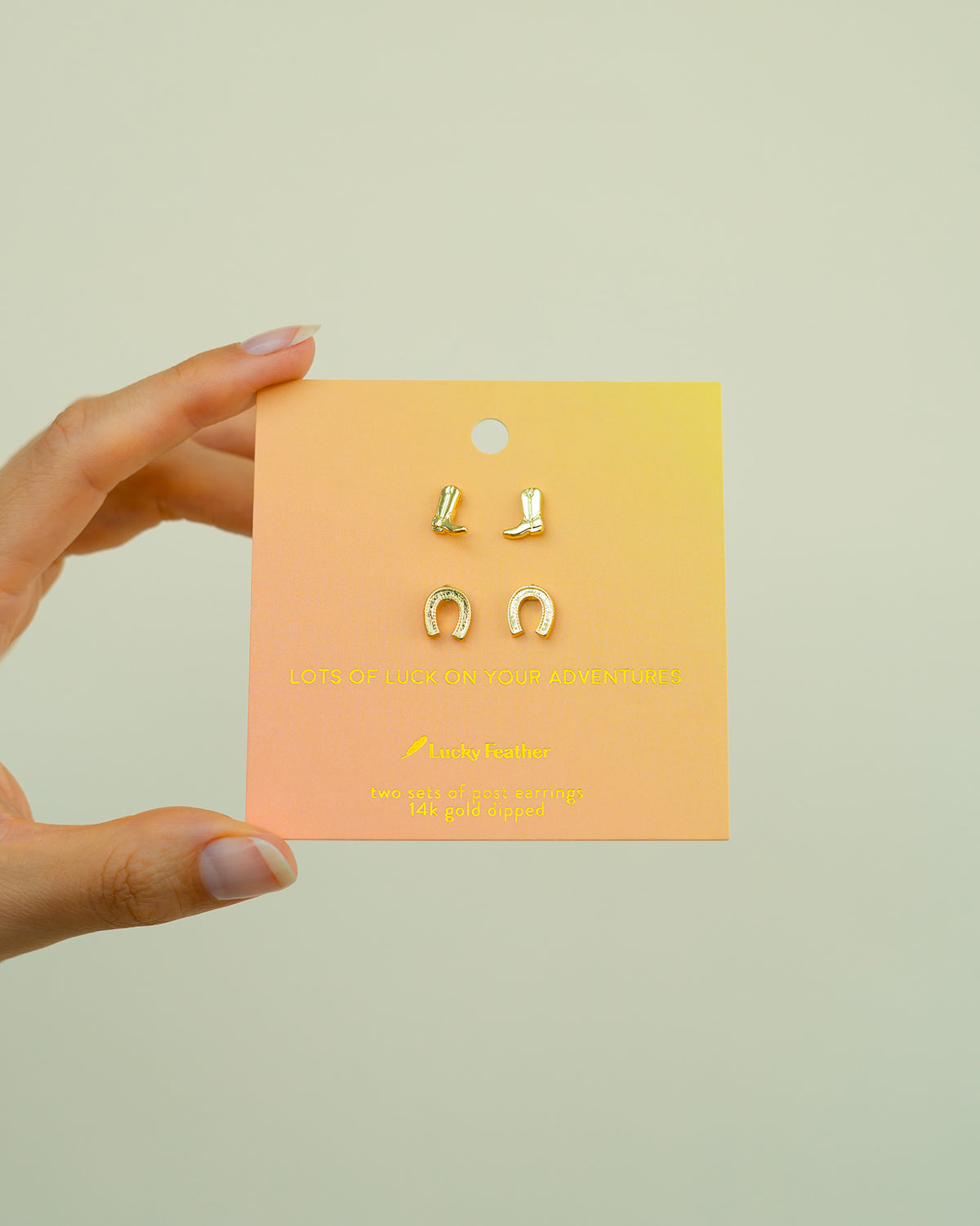 A hand holds a card displaying gold-colored horseshoe and cowboy boot earrings.