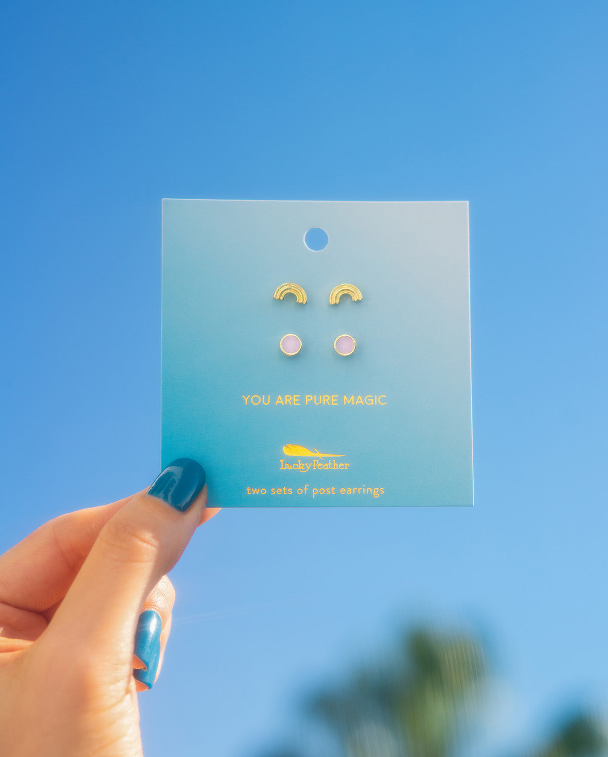 A hand holding a Lucky Feather earring card with two sets of post earrings against a blue sky.