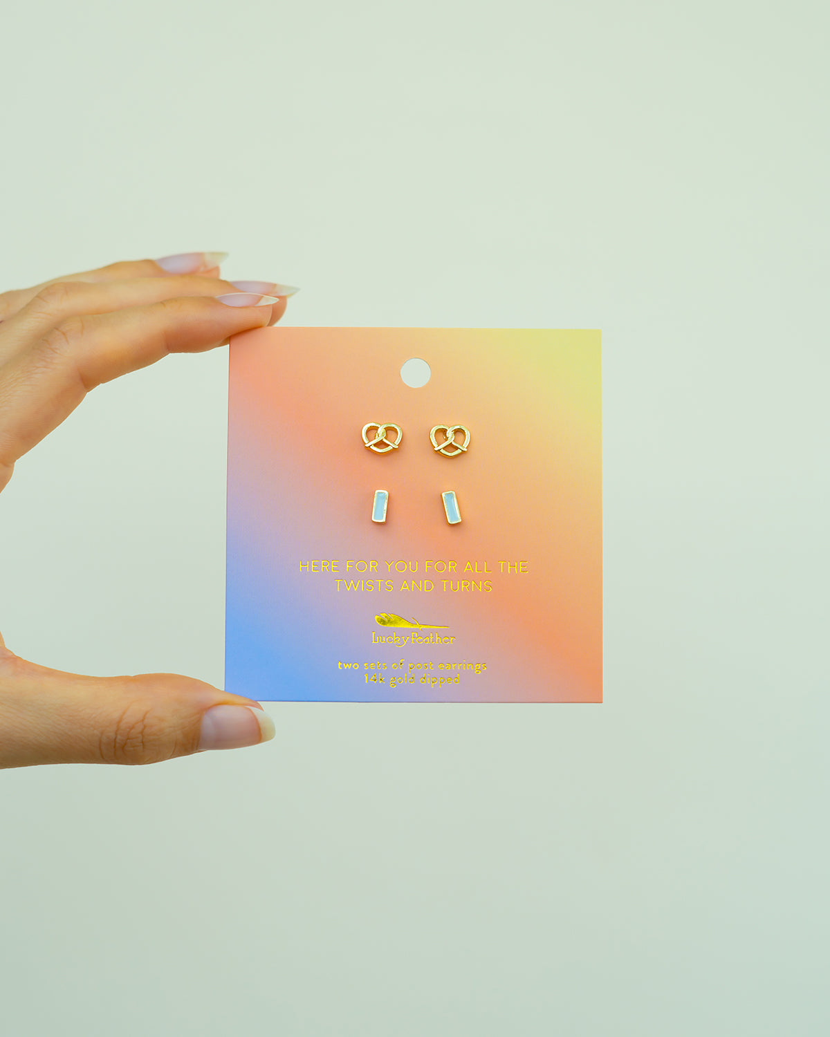 A hand holds a card displaying two sets of earrings: pretzel-shaped and rectangular, labeled as Lucky Feather 14k gold dipped.