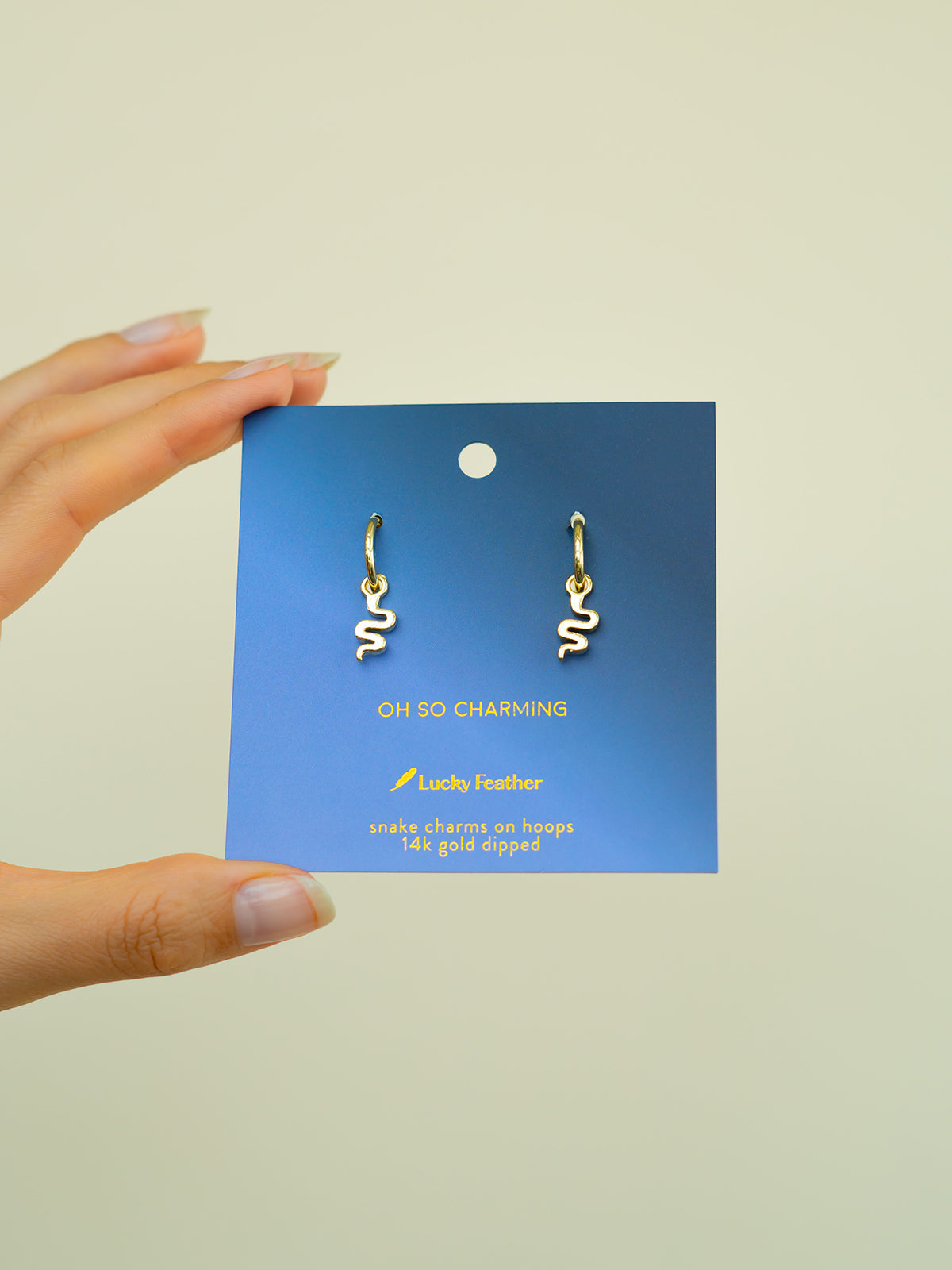 A hand holds a blue card displaying a pair of gold hoop earrings with snake charms.