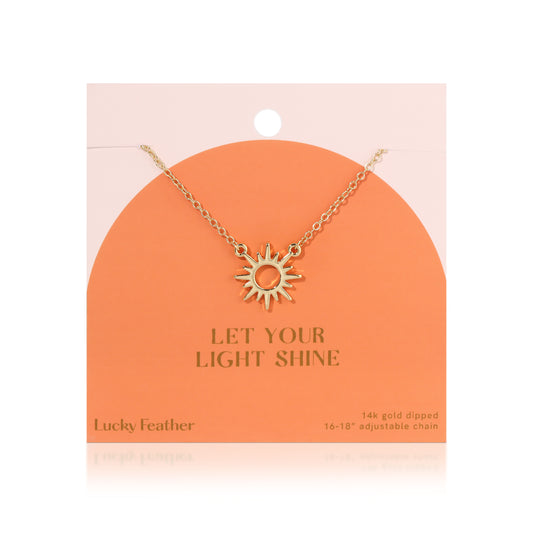 A gold-colored sun pendant hangs on a chain from a peach-colored display card that reads 'Let Your Light Shine'.