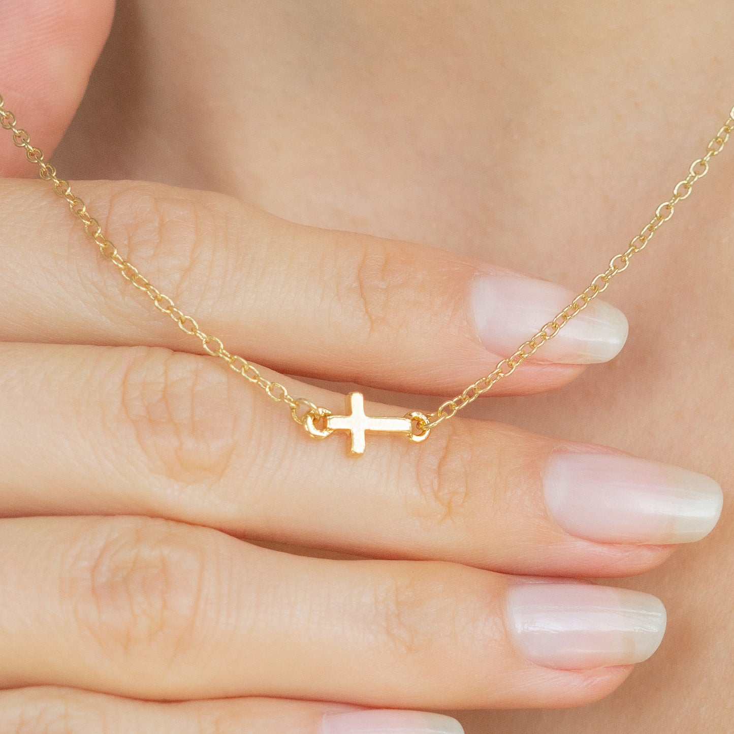 A hand holds a gold chain necklace with a sideways cross pendant.