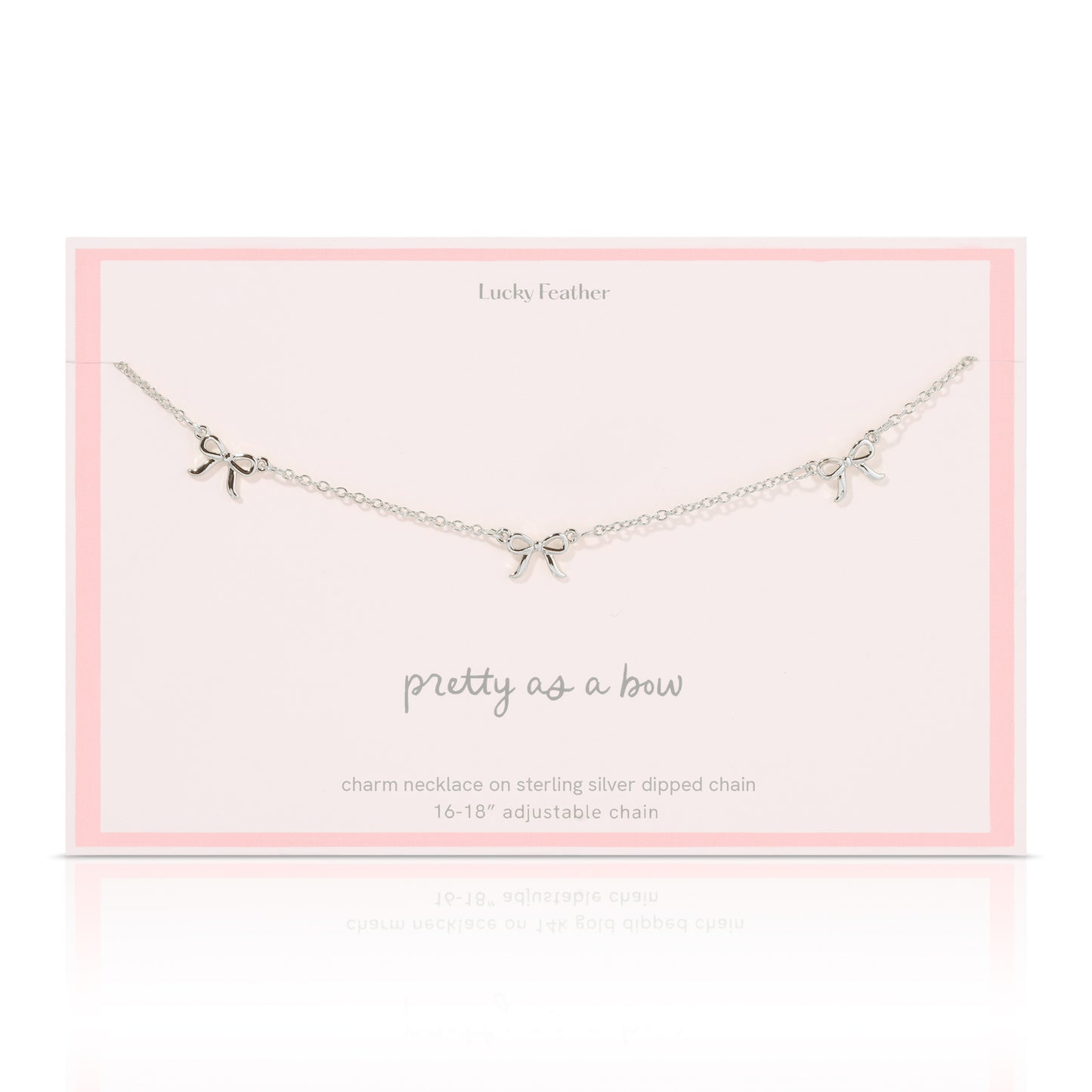 A silver-colored necklace with three bow charms lies on a pale pink card from "Lucky Feather".
