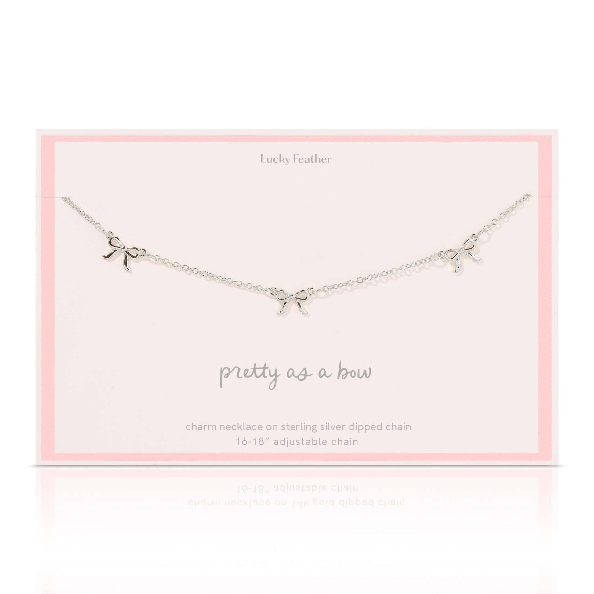A silver-colored necklace with three bow charms lies on a pale pink card from "Lucky Feather".