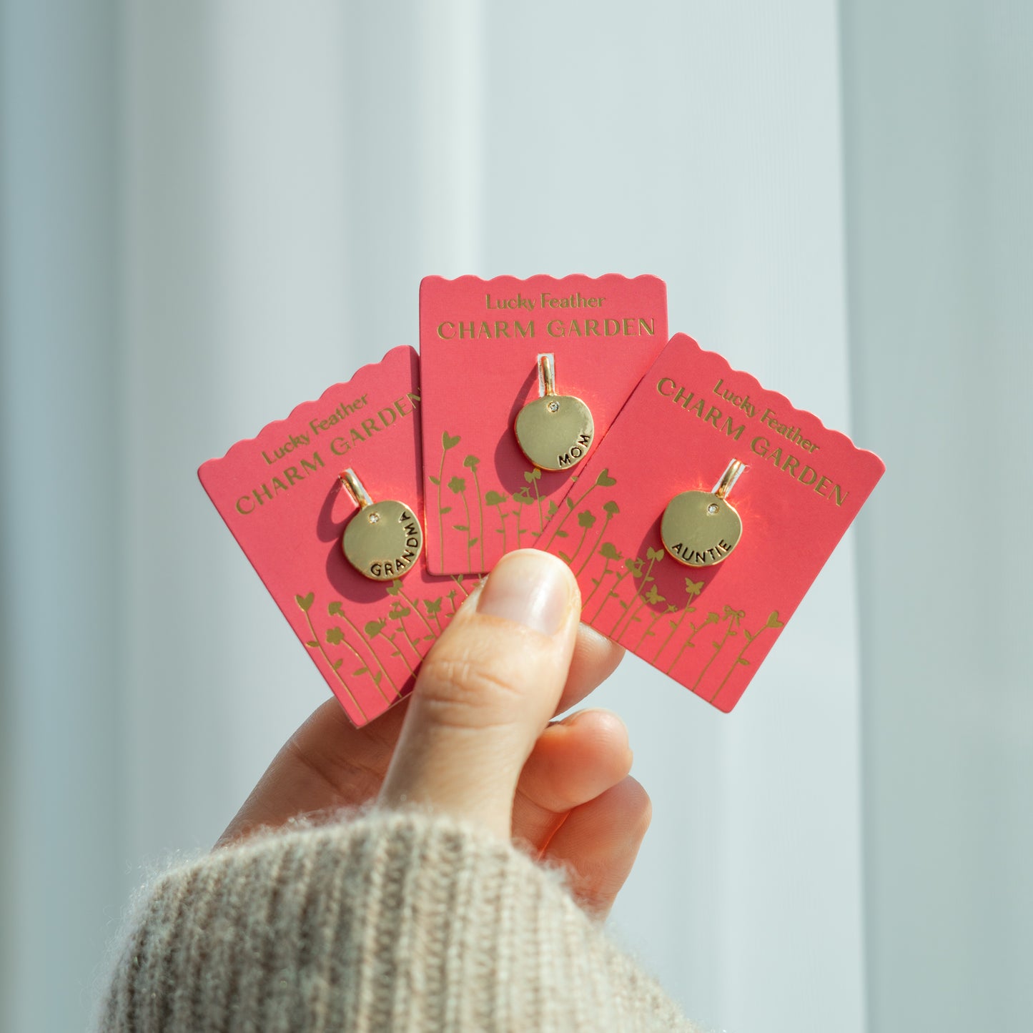 A hand holds three "Lucky Feather Charm Garden" pendants with the words "Grandma", "Mom", and "Auntie".