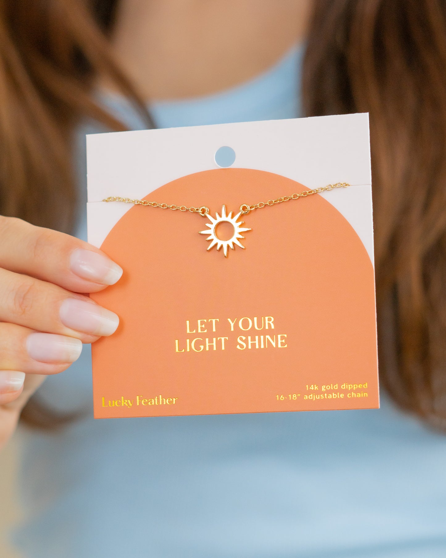 A hand holds a product card displaying a gold-colored necklace with a sun pendant.