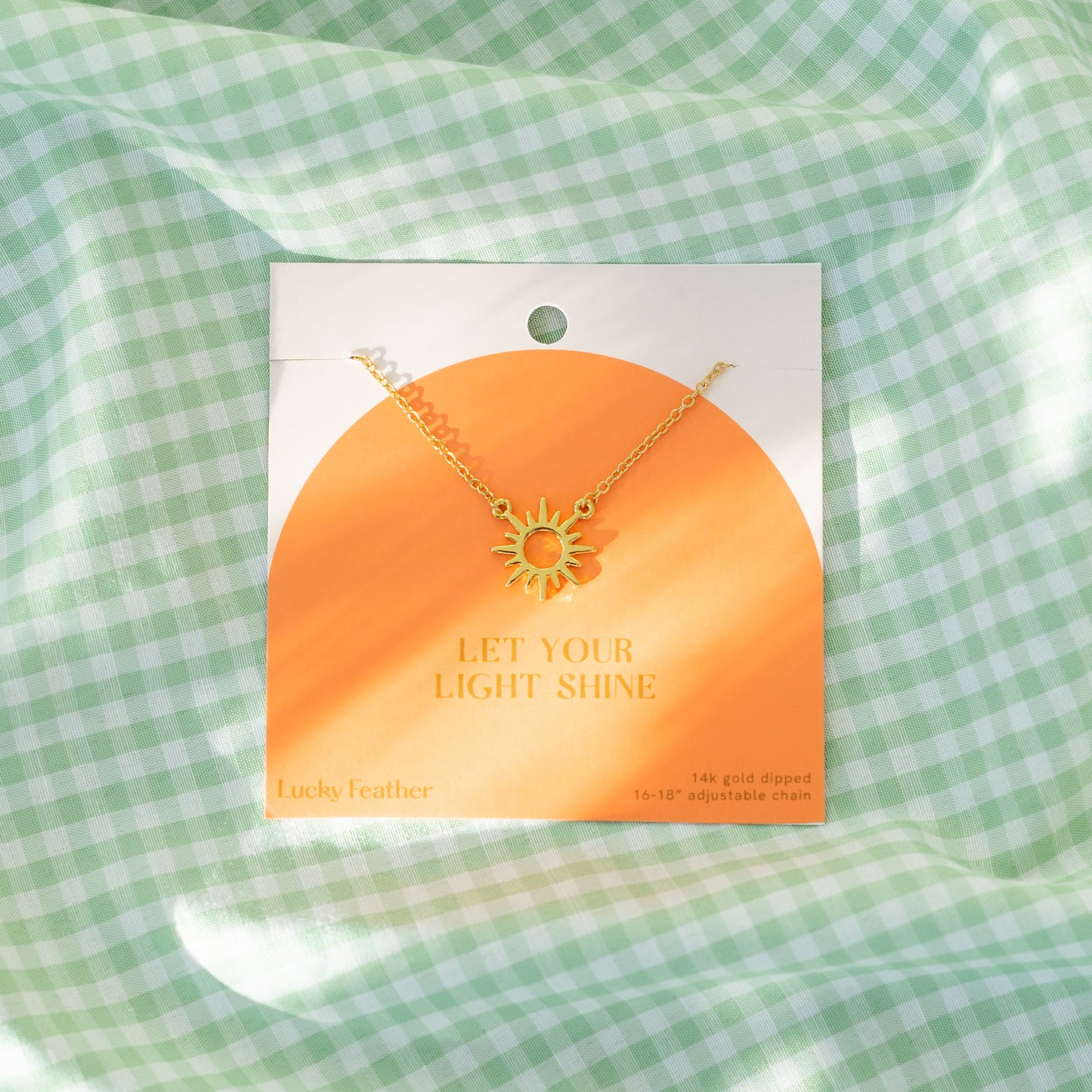 A gold-colored sun pendant necklace on a card that reads "Let Your Light Shine," displayed on a light green and white checkered fabric.