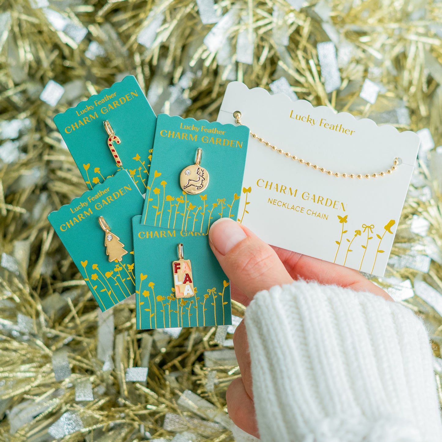 A hand in a white sweater holds several Lucky Feather Charm Garden charms and a necklace chain against a background of gold and silver tinsel.