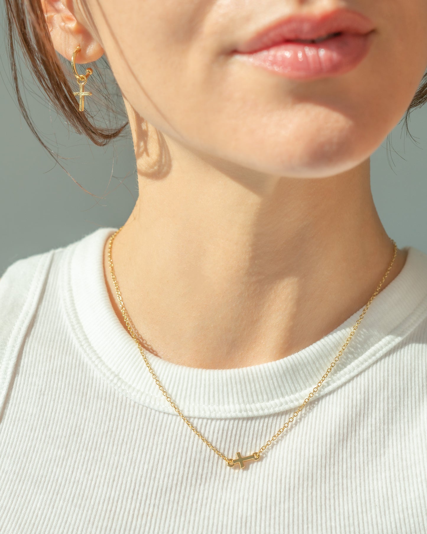 A woman wears gold cross jewelry, including a necklace and earrings, with a white ribbed tank top.
