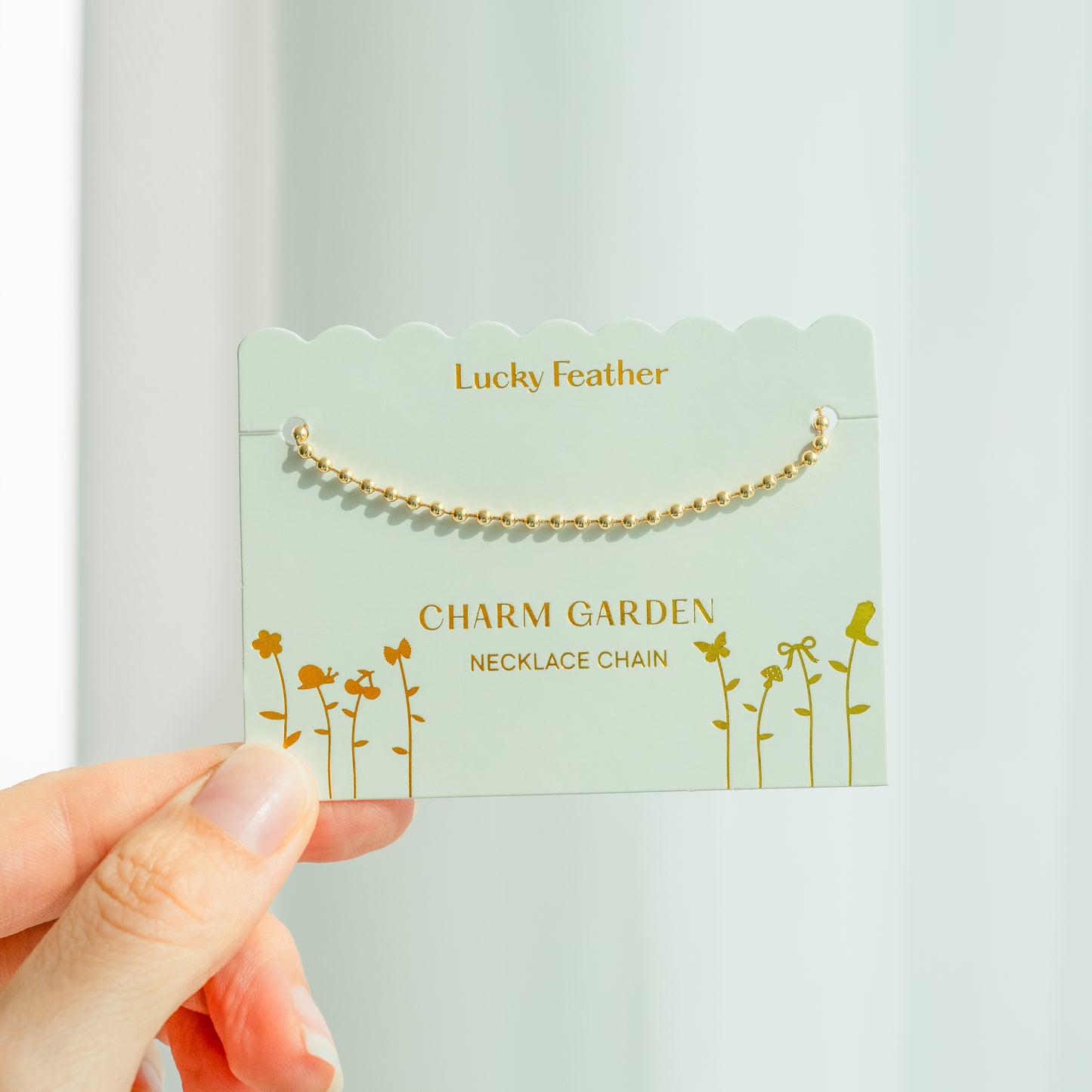 A hand holds a "Lucky Feather Charm Garden Necklace Chain" product card, with a golden ball chain necklace attached.
