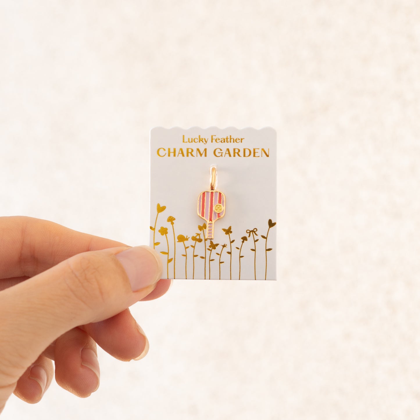 A hand holds a "Lucky Feather Charm Garden" card with a pickleball paddle charm attached.