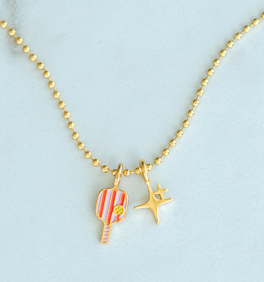 Close-up of a gold beaded necklace with a pickleball paddle charm and a Mid-Century Modern starburst charm.