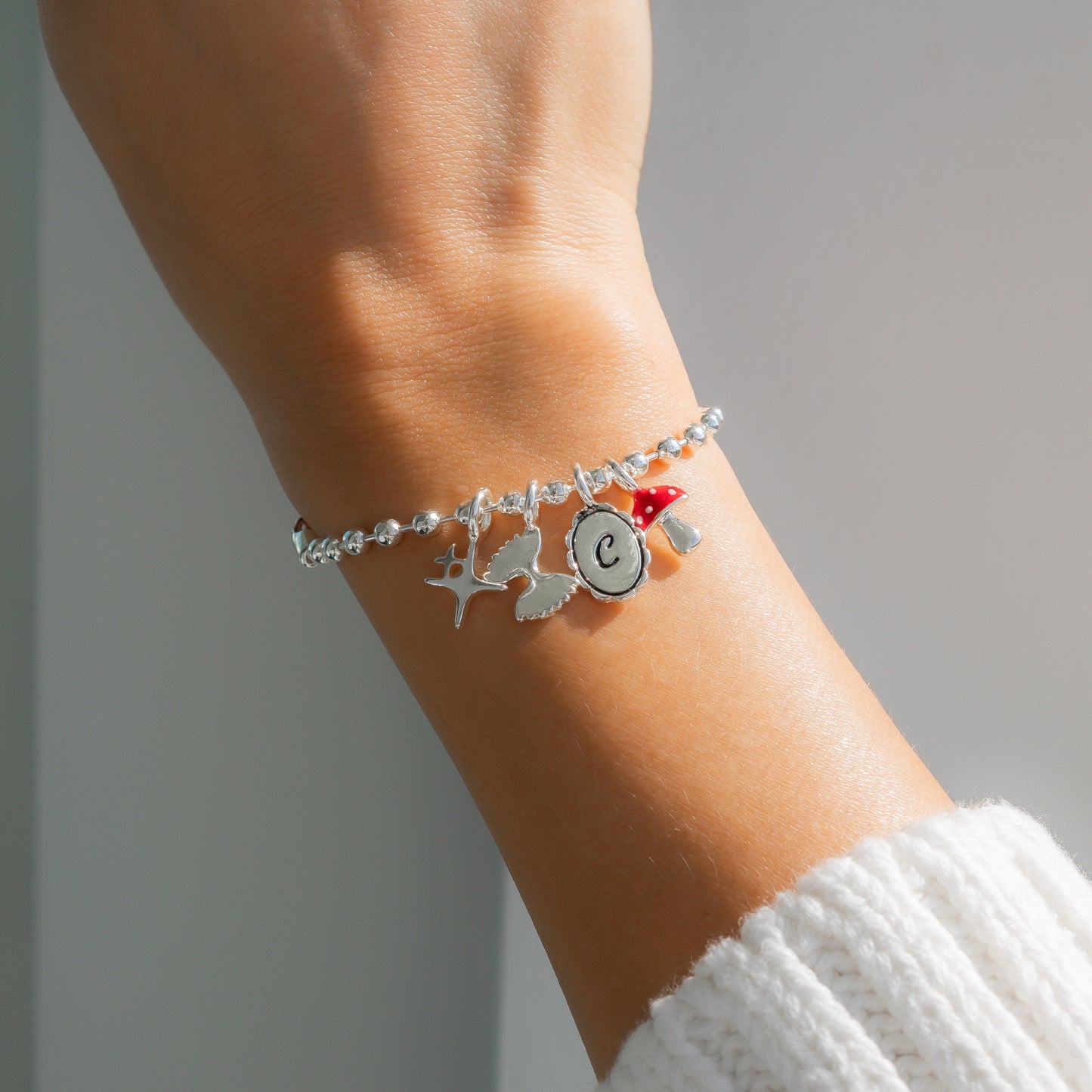 A silver charm bracelet with a letter "C" and a mushroom charm is on a wrist.