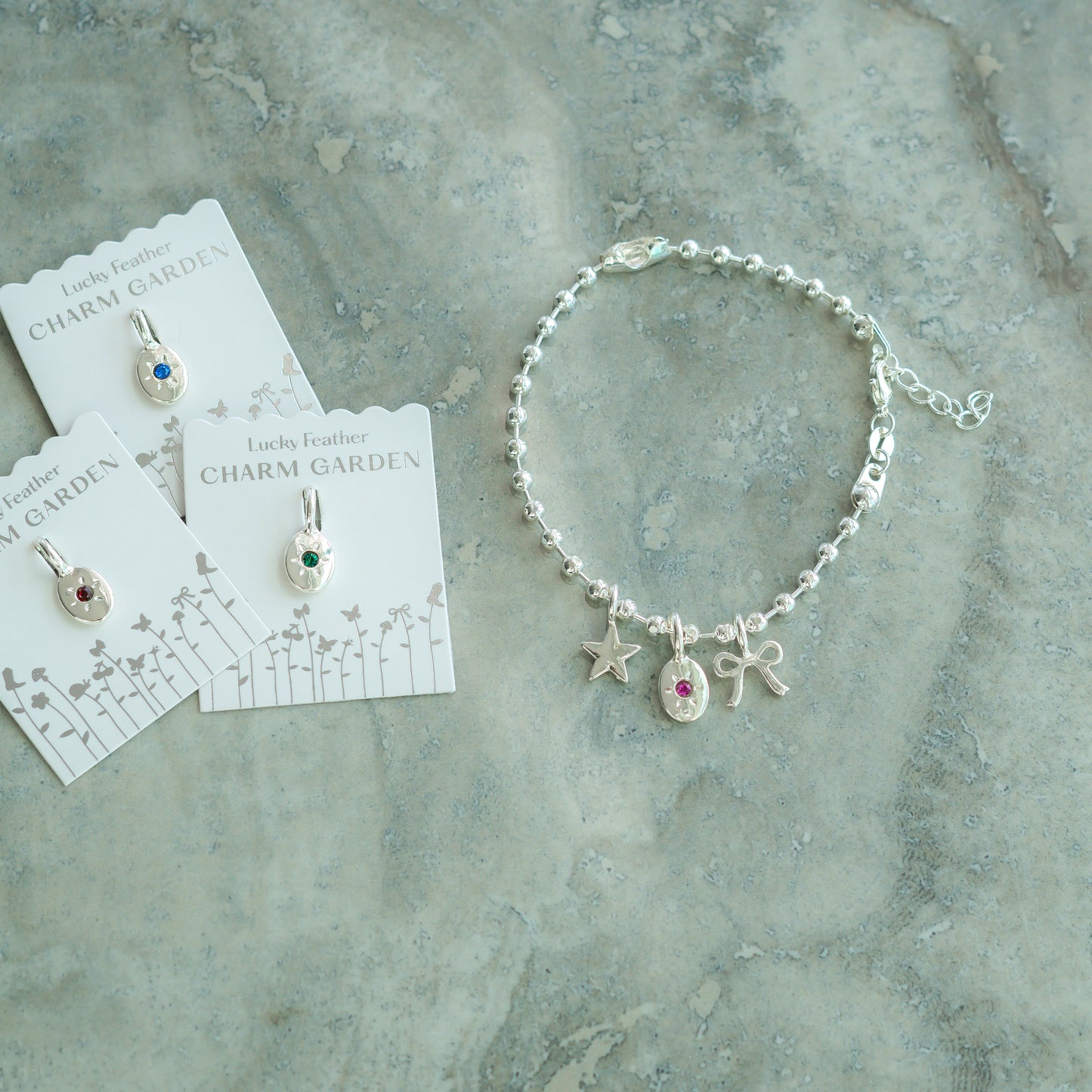A silver charm bracelet and charm pendants on cards are arranged on a textured gray surface.