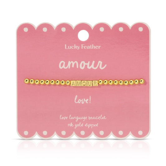 A gold beaded bracelet spelling 'AMOUR' sits on a pink scalloped display card.