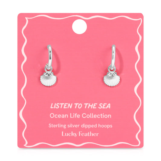 Two sterling silver hoop earrings with seashell charms are displayed on a pink card.