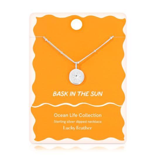 A silver 'Bask in the Sun' necklace hangs on a bright orange and white display card.