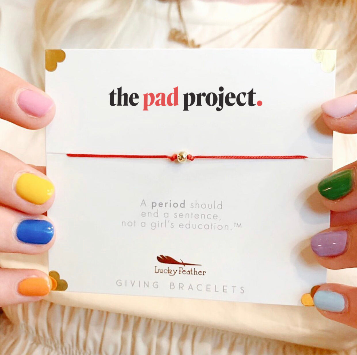 Hands with multicolored nails hold a card with a red string bracelet, titled "the pad project," and the text "A period should end a sentence, not a girl's education."