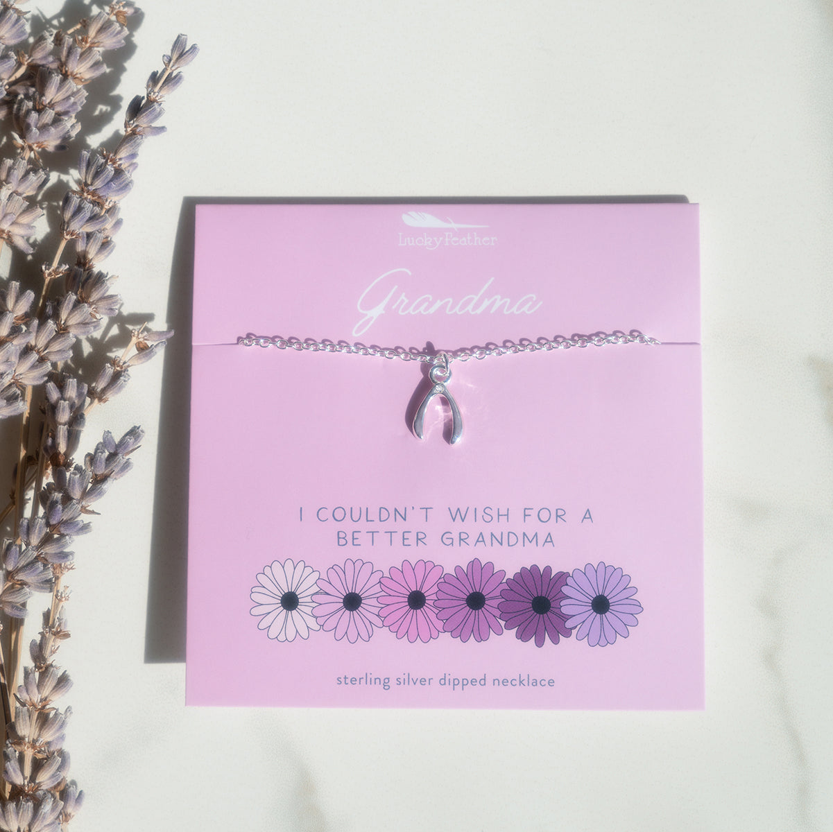 A silver necklace with a wishbone charm rests on a pink card with the text "Grandma" and an arrangement of daisies, next to dried lavender.