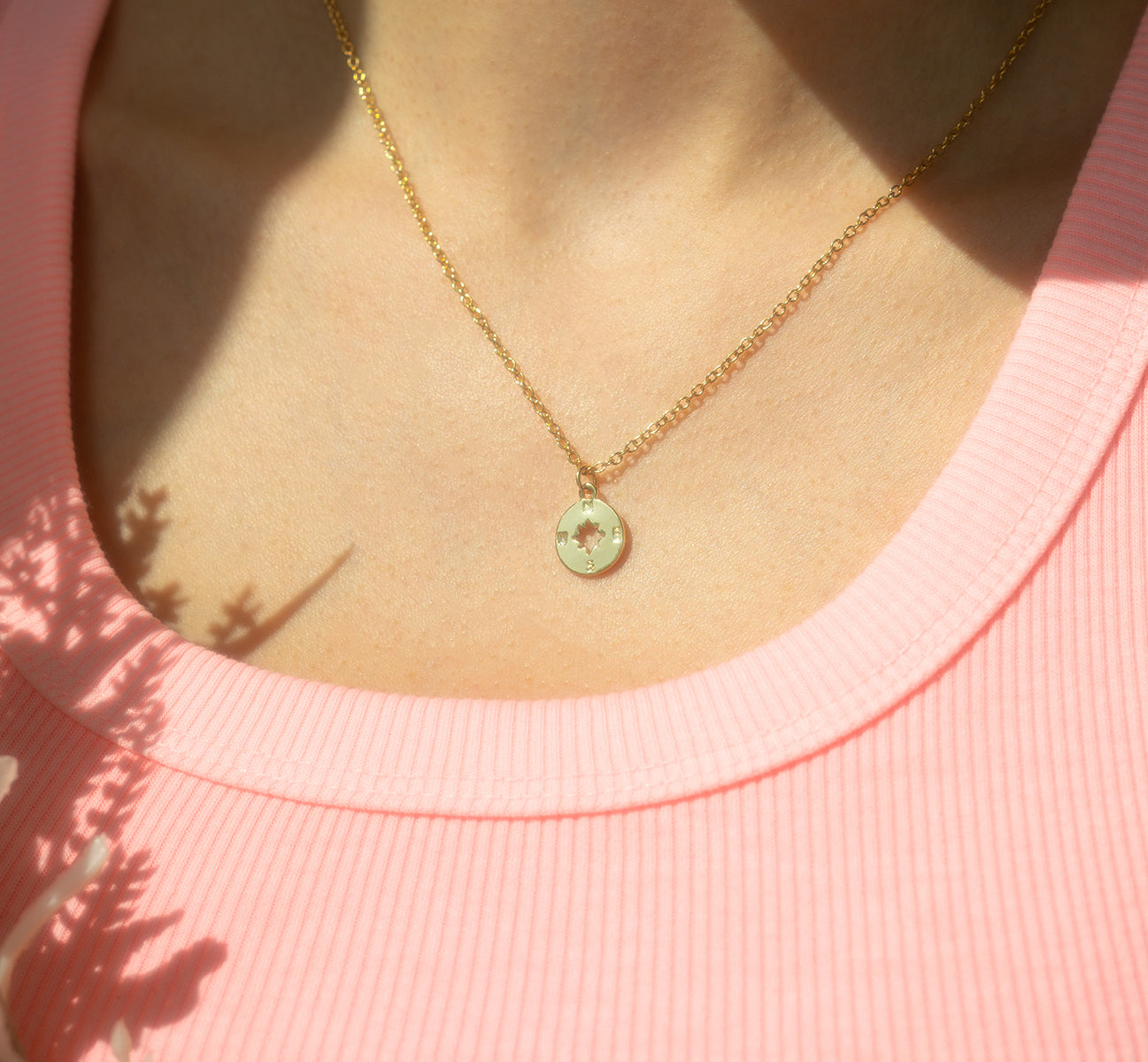 A gold chain necklace with a compass charm is worn with a pink shirt.