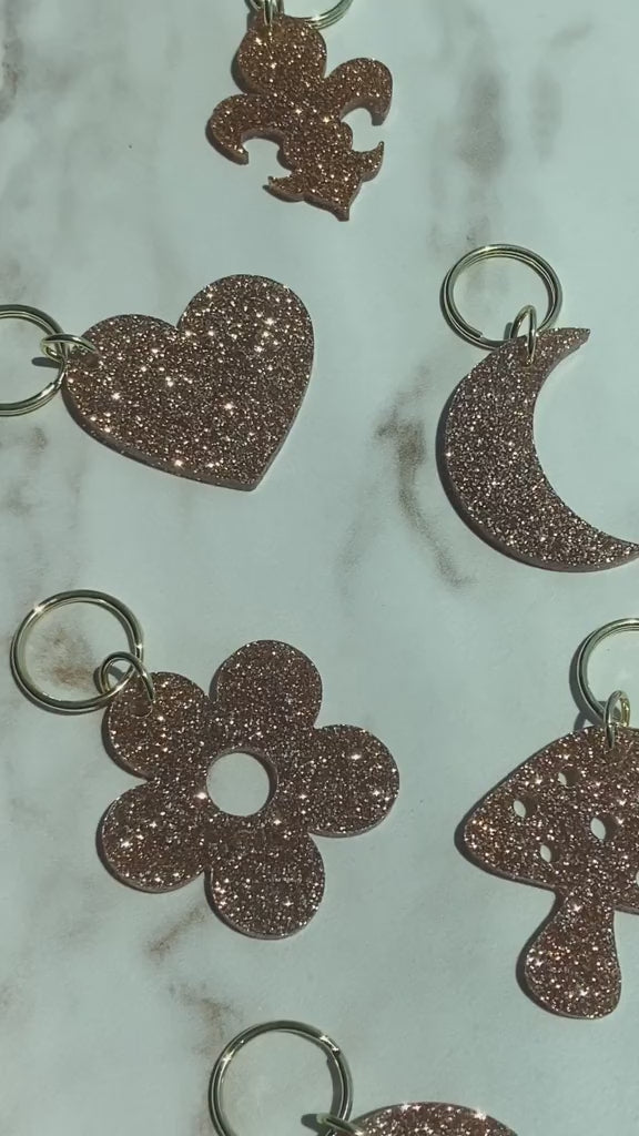 Close-up of several rose-gold glittery keychains in shapes of a fleur-de-lis, heart, crescent moon, flower, and mushroom on a white marble surface.