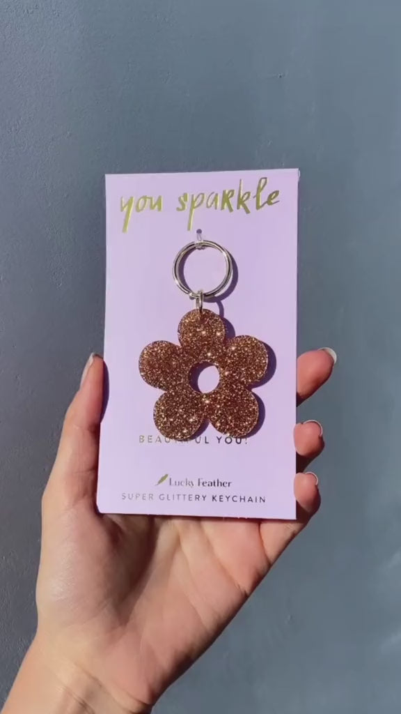 A hand holds a lilac card with the words 'you sparkle' and a flower-shaped glittery keychain.