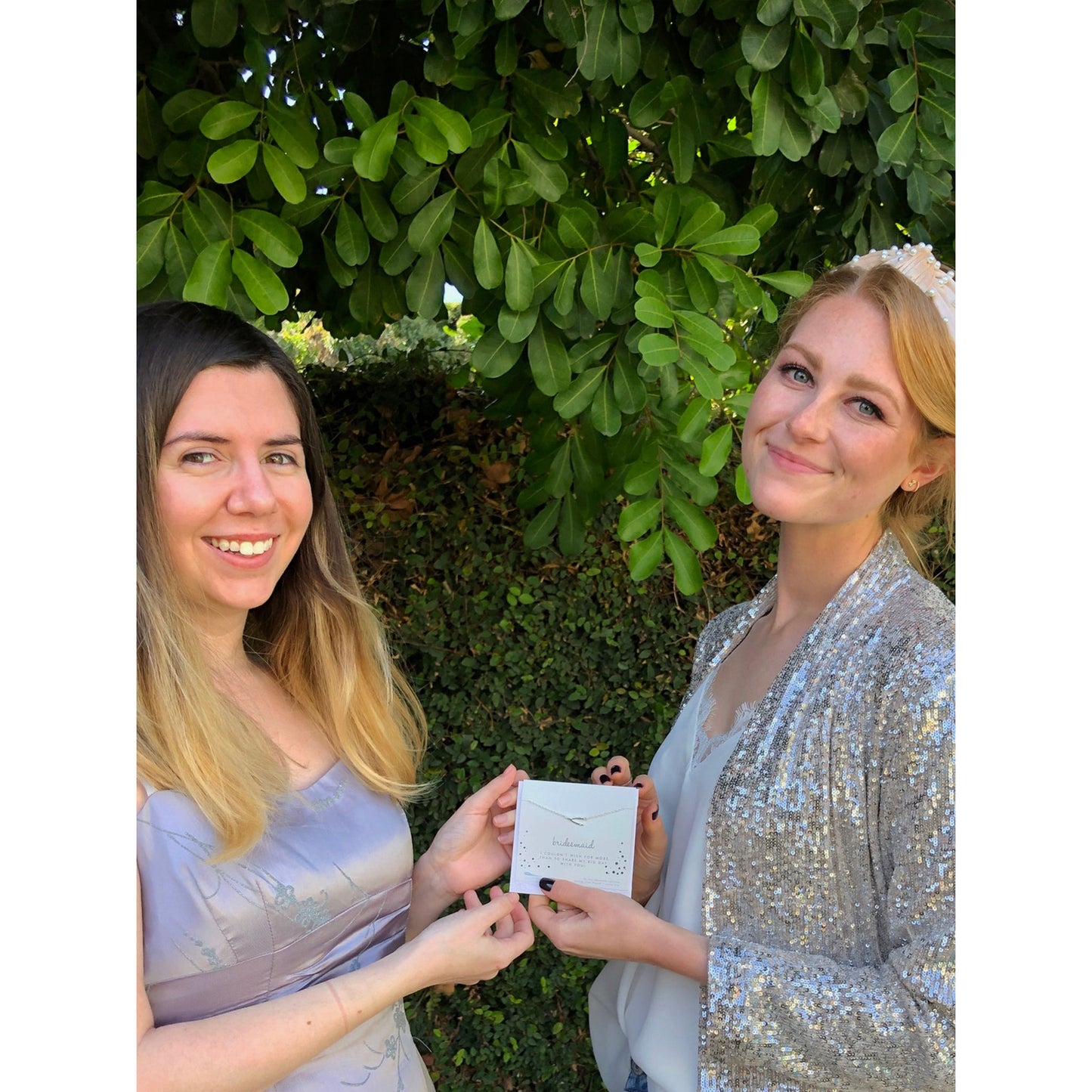 Two women pose in front of foliage, holding a gift card that reads "bridesmaid."