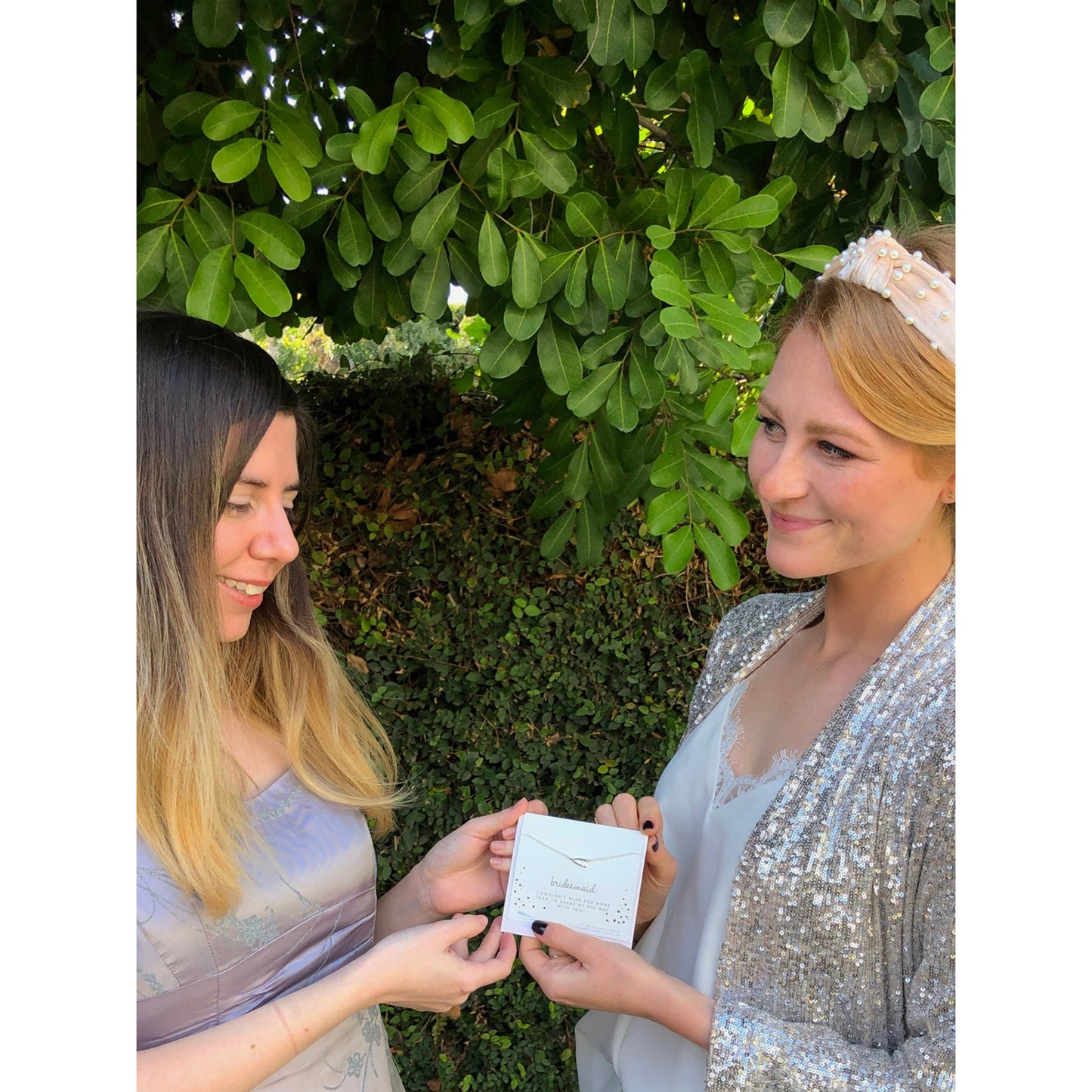 Two women stand outdoors in front of greenery, one handing the other a card that reads "bridesmaid."