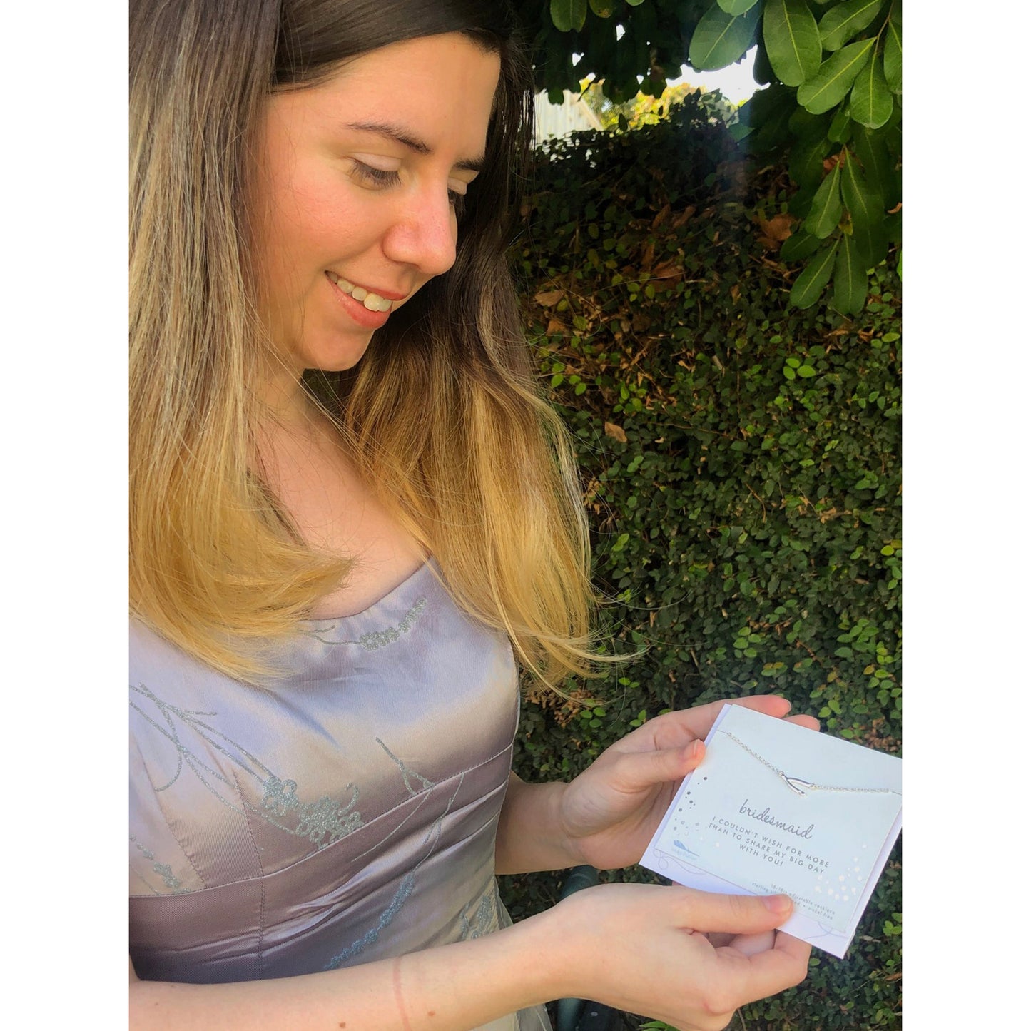 A woman in a dress is smiling and holding a card that reads "bridesmaid" with a necklace attached.