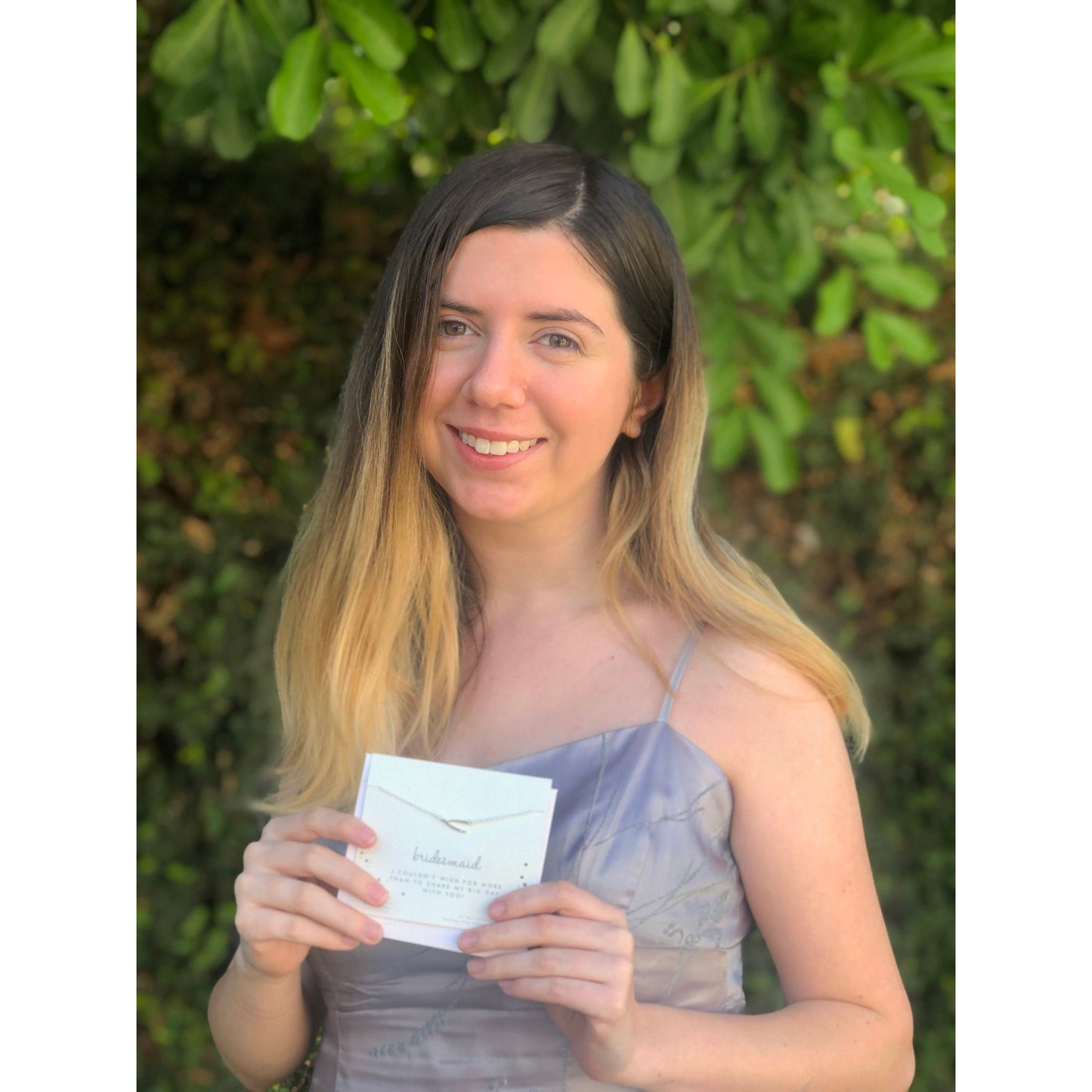 A woman in a dress smiles and holds a card that says "bridesmaid" with a necklace attached.