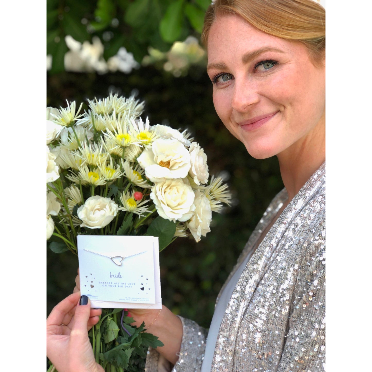 A smiling woman in a sequined jacket holds a bouquet of flowers and a "Bride" necklace card.