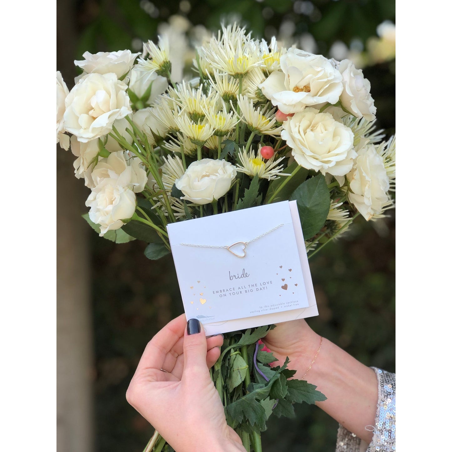 A person holding a bouquet of white flowers with a "bride" heart necklace in front.