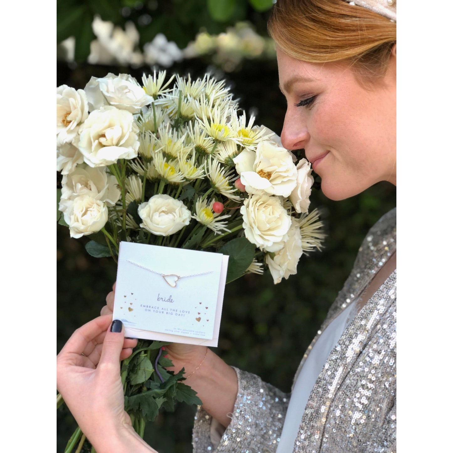A woman smelling a bouquet of white roses and daisies holds a card featuring a necklace that reads "bride."
