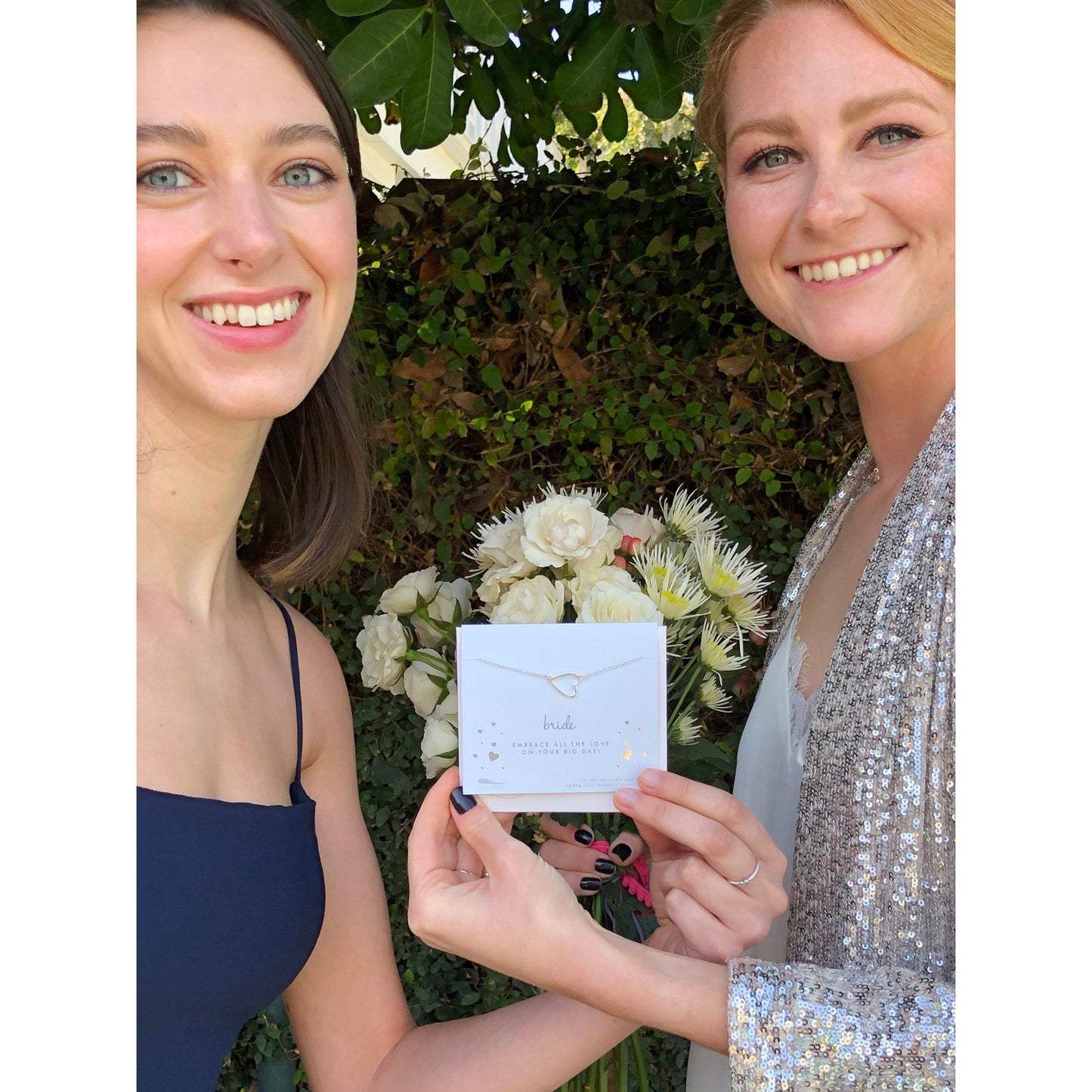 Two smiling women hold a "bride" necklace in front of a bouquet of flowers and a green leafy background.