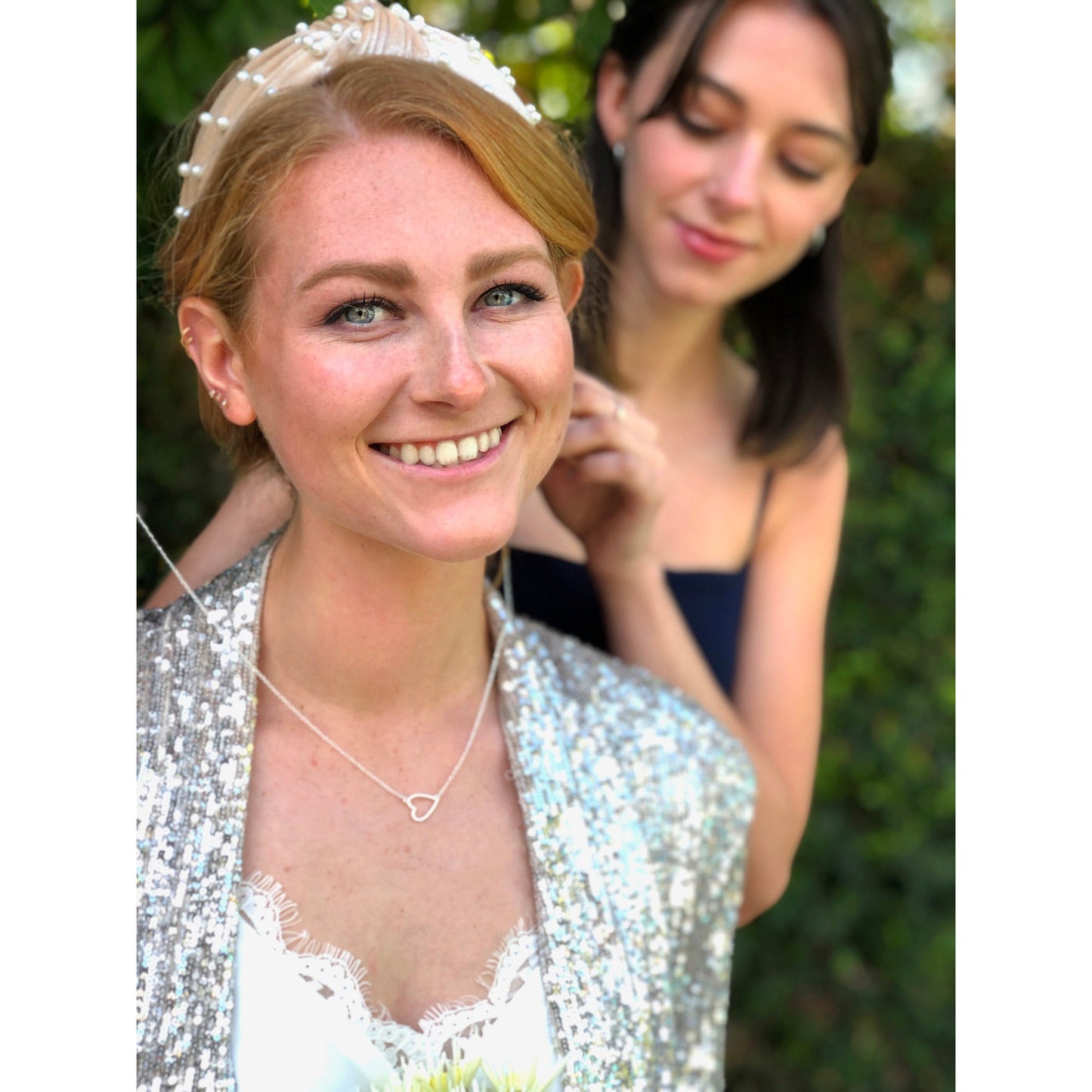 A smiling woman with a beaded headband and heart necklace has a second woman adjusting her necklace.