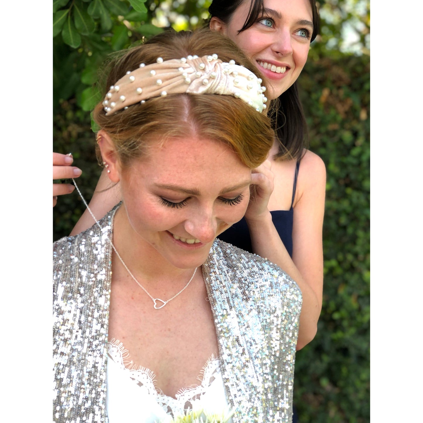A woman smiles as another woman puts a necklace on her; she is wearing a sequined jacket and a headband with pearls.