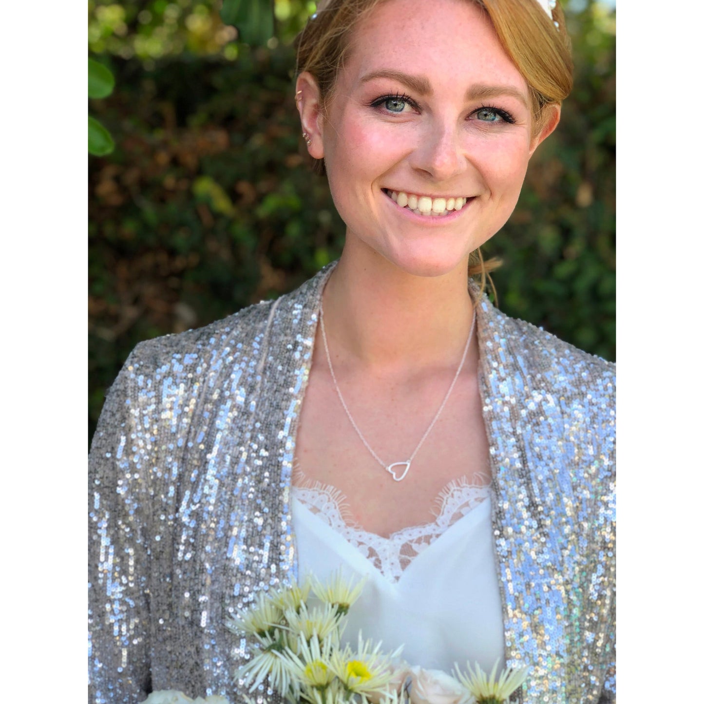 A smiling woman wearing a sequined jacket and heart necklace holds flowers.